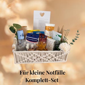 May include: A woven basket filled with various items, including a blue deodorant, bandages, creams, and small gifts. The basket is decorated with greenery and a white flower. Text at the bottom reads "For small emergencies Complete Set".