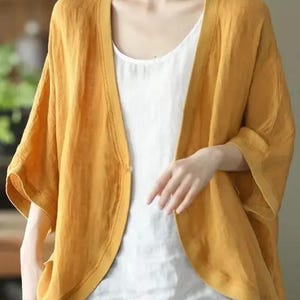 May include: Mustard yellow open-front cardigan with elbow-length sleeves. The cardigan is worn over a white tank top and paired with blue pants. The cardigan has a relaxed fit and a simple design, suitable for casual wear.