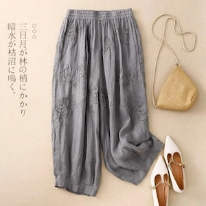 May include: Grey linen trousers with floral embroidery, a straw handbag, and white flats with gold chain detailing. The trousers have an elasticated waistband and a wide leg. Japanese text is visible on the left.