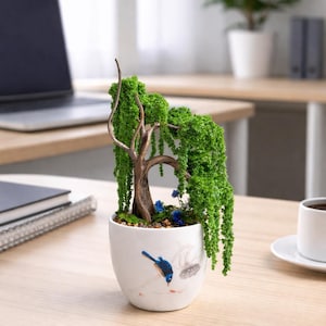 Op de afbeelding: Een decoratieve kunstmatige wilgenboom met levendig groen blad en een bruine, getextureerde stam, geplant in een witte keramische kom. De kom is voorzien van een geschilderd motief van een blauwe vogel en bloemen. De basis is gevuld met steentjes en mos.