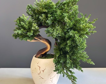 May include: A faux bonsai tree with dense, vibrant green needle-like foliage and a curved, dark brown trunk. The tree is planted in a rounded, off-white ceramic pot featuring a decorative cracked texture across its exterior surface.