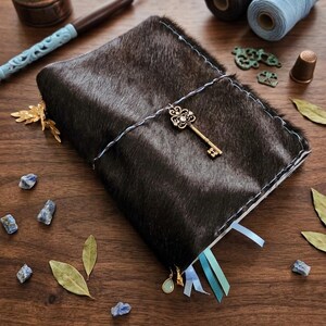 May include: A dark brown leather journal with a decorative key charm, blue and white stitching, and blue ribbon bookmarks. The journal is surrounded by writing supplies, including an inkwell, pen, and spools of thread, on a wooden surface.
