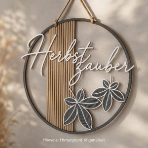 Könnte beinhalten: Ein kreisförmiges Wandobjekt mit einem dunklen Rahmen, einem vertikalen Paneel aus hellbraunen Lamellen und dem weißen Schriftzug "Herbstzauber". Zwei dunkelgraue Blattausschnitte mit weißen Umrandungen sind unten rechts angebracht.