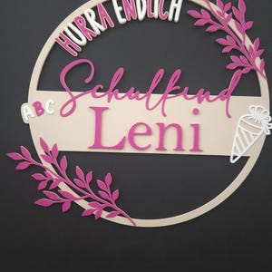 Könnte beinhalten: Ein rundes Wandschild aus Holz mit dem Schriftzug Hurra Endlich Schulkind Leni in Pink und Weiß. Das Design umfasst pinkfarbene Blattzweige an den Seiten, einen kleinen weißen ABC-Block und eine weiße Schultüte auf einem hellbeigen Holzrahmen.