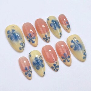 Puede incluir: Un conjunto de diez u&ntilde;as postizas en forma de almendra. Las u&ntilde;as presentan un dise&ntilde;o con una base amarilla clara, detalles florales azules y una punta color melocot&oacute;n. El dise&ntilde;o floral est&aacute; pintado a mano.
