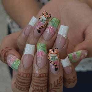 May include: A set of long, sculpted fingernails with a tiger theme. The nails feature a light pink base, white tips, and 3D tiger figurines. Floral and leaf designs are painted on some nails, with water droplets.