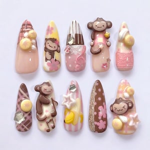 Op de afbeelding: Een verzameling van tien amandelvormige nail art ontwerpen. De nagels hebben verschillende ontwerpen, waaronder apenfiguren, muzieknoten en bloempatronen. Kleuren zijn onder meer roze, bruin, geel en wit, met decoratieve versieringen.
