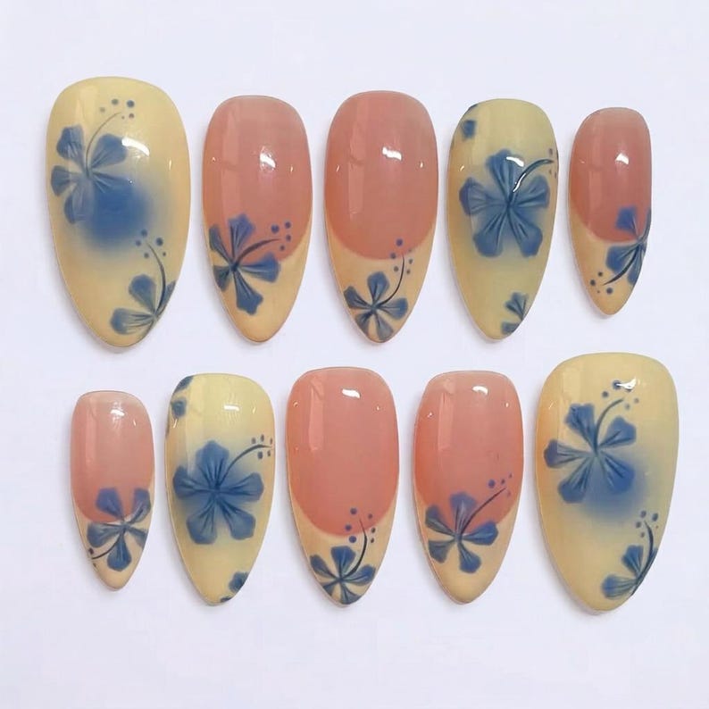 Puede incluir: Un conjunto de diez u&ntilde;as postizas en forma de almendra. Las u&ntilde;as presentan un dise&ntilde;o con una base amarilla clara, un estampado floral azul y un acento rosa. El dise&ntilde;o floral es una flor de hibisco azul con hojas azul oscuro.