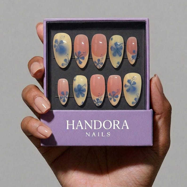 Puede incluir: Un juego de U&Ntilde;AS HANDORA en una caja morada. Las u&ntilde;as artificiales son de forma ovalada con una base amarilla clara, puntas rosas y dise&ntilde;os florales azules. Las u&ntilde;as se sostienen en una mano.