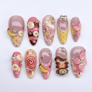 Könnte beinhalten: Zehn künstliche Fingernägel mit unterschiedlichen Designs und Dekorationen. Die Nägel zeigen eine Palette von Rosa, Gelb und Braun, verziert mit Perlen, Blumenmustern und Cartoonfiguren. Einige Nägel haben ein Bananenmotiv und bieten eine verspielte Ästhetik.