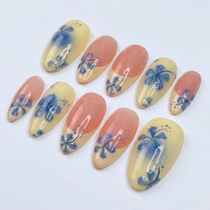 Puede incluir: Un conjunto de diez u&ntilde;as artificiales de varios tama&ntilde;os. Las u&ntilde;as presentan una base amarillo claro, un dise&ntilde;o floral azul y una punta color melocot&oacute;n. Las u&ntilde;as ovaladas est&aacute;n dispuestas sobre una superficie blanca.