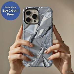 Puede incluir: Funda de teléfono plateada con un diseño de papel de aluminio arrugado, sostenida frente a un fondo neutro. La funda tiene un brillo metálico y una apariencia texturizada. Un círculo azul con el texto "Bundle & Save Buy 2 Get 1 Free" está en la esquina superior izquierda.