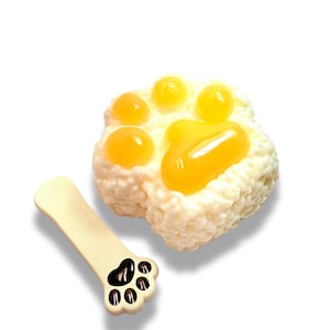 May include: A whimsical, off-white paw-shaped item with yellow accents and a bone-shaped accessory. The paw has five yellow circles and a larger yellow oval, resembling a fried egg. The bone has a paw print with black details.
