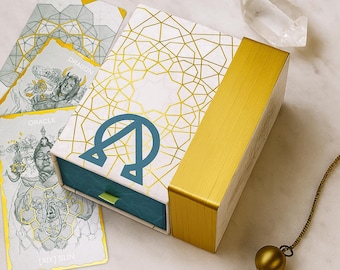 May include: A rectangular oracle deck box with a white and gold geometric pattern, a blue omega symbol, and a gold side panel. Beside the box are two oracle cards labeled Dragon and Sun, a clear quartz crystal, and a gold-toned metal pendulum.