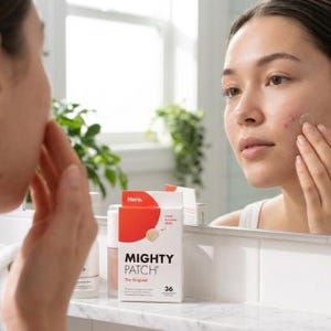 May include: A woman applying a Mighty Patch acne patch to her face in front of a mirror. The box of Mighty Patch is white and orange, with the text "MIGHTY PATCH" and "The Original". The box contains 36 patches.