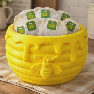 May include: A bright yellow, bee-themed tea bag holder. The holder is shaped like a beehive with dripping honey accents and a sculpted bee. The container is filled with tea bags.