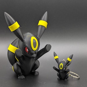 May include: A black and yellow Umbreon figurine and a matching smaller Umbreon keychain. Both figures feature pointed ears with yellow bands, a yellow oval on the forehead, and red eyes. The larger figure is in a seated pose, while the keychain has a ring.