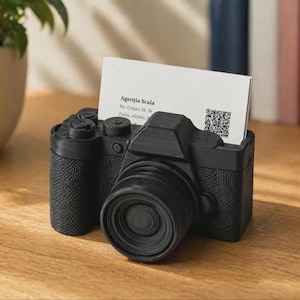 May include: A matte black camera-shaped business card holder with a textured grip surface. A white rectangular business card is inserted into the top slot, displaying the text Agentia Scala, Str. Crisan, bl. 36, Zalau, 450061, and a QR code.