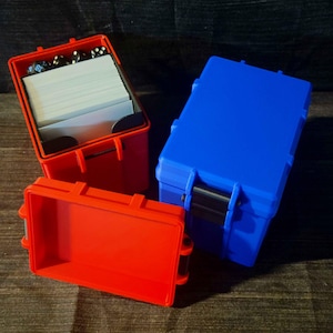 May include: Three colorful, rectangular storage cases. One red case is open, revealing white cards and dice. A second red case is empty. A blue case is closed. All cases have black latches.
