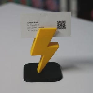 May include: A yellow lightning bolt-shaped business card holder with a black base. The business card holder is holding a white business card with the text "Agenția Scala" and contact information.