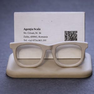 May include: A beige eyeglasses-shaped business card holder with a card displaying the text "Agenția Scala" and contact information. The holder is made of a solid material and sits on a gray surface.