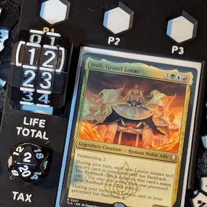 May include: A black game board with a Magic: The Gathering card, "Iroh, Grand Lotus." Includes a life counter, a tax die, and player markers. The card is gold and green with a detailed illustration.