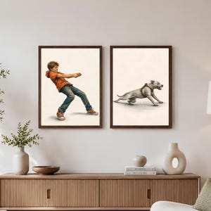 May include: Two framed wall art prints featuring a tug-of-war scene. The left print shows a person in an orange hoodie and blue jeans pulling a rope. The right print shows a light-colored dog wearing a brown harness pulling in the opposite direction.