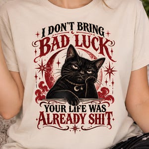 May include: A cream-coloured t-shirt featuring a graphic of a black cat with a crescent moon collar. The text reads I DON'T BRING BAD LUCK and YOUR LIFE WAS ALREADY SHIT. The design includes a red moon, stars, and swirling clouds in a dark red ink.