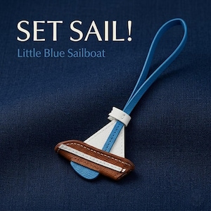 Könnte beinhalten: Ein Segelboot-Schlüsselanhänger in Blau und Weiß mit braunem Boden und blauem Riemen. Der Text "SET SAIL!" und "Little Blue Sailboat" sind in Weiß über dem Schlüsselanhänger auf einem dunkelblauen Hintergrund zu sehen.