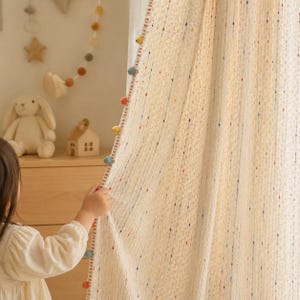May include: A cream-colored curtain featuring a textured waffle-weave fabric with vertical rows of small, multicolored dots. The edge of the curtain is finished with a decorative trim adorned with a series of small, round, multi-colored pom-poms.
