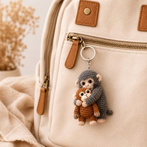 May include: A crochet keychain featuring a large grey monkey hugging a smaller brown monkey. The monkeys have embroidered facial features and are attached to a silver metal keyring. The keychain is clipped to the zipper of a beige canvas backpack.