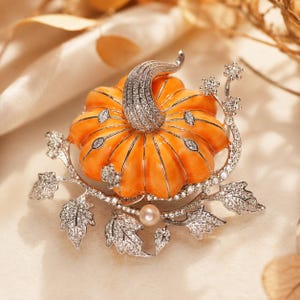 May include: A pumpkin-shaped brooch featuring a vibrant orange enamel body with a silver-toned stem encrusted with clear crystals. The design includes silver-toned leaves and vines accented with clear crystals and a single small white pearl.