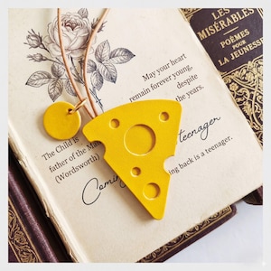 Puede incluir: Un colgante amarillo con forma de queso con un dije redondo, ambos en un cordón marrón. El colgante tiene tres agujeros pequeños y un agujero central grande. El colgante está sobre un libro abierto con texto.