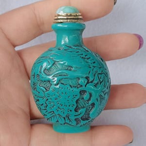 May include: A small, round, turquoise-colored snuff bottle featuring intricate, deep-relief carvings of a dragon and floral patterns. The bottle is topped with a gold-toned metal cap featuring a smooth, light blue, rounded stone inlay at the very top.