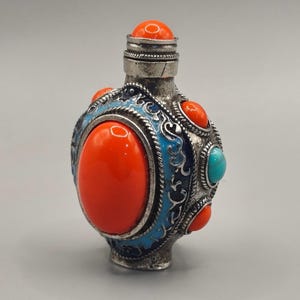 May include: A small, oval-shaped metal snuff bottle featuring a large, smooth, bright orange cabochon stone at the center. The bottle is decorated with blue enamel filigree, smaller orange stones, and a single turquoise stone on the side.