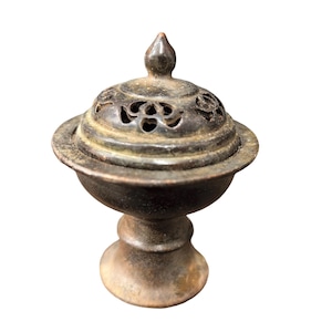 May include: A dark, weathered metal incense burner featuring a pedestal base, a wide bowl, and a removable lid with an intricate cutout pattern and a pointed finial. The surface shows a matte, aged patina with subtle brown and black tones.
