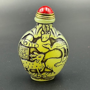 May include: A pale yellow snuff bottle featuring black etched illustrations of deer and birds. The bottle has a narrow neck with a metallic gold-toned rim and a rounded, bright red stopper cap. The surface displays detailed animal carvings.