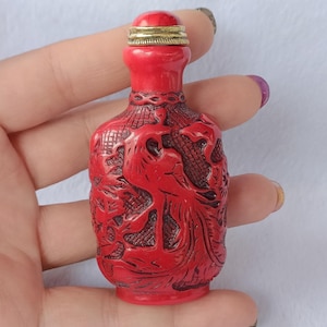Puede incluir: Una pequeña botella de rapé roja con intrincados relieves tallados de un pájaro y motivos florales. La botella tiene un cuello estrecho y un tapón redondeado con una banda metálica dorada. La superficie presenta un acabado rojo mate texturizado.