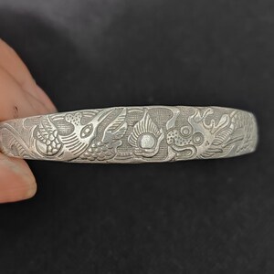 May include: A silver-toned metal bangle bracelet featuring an intricate embossed design of a mythical dragon and a phoenix facing each other, separated by a central flaming pearl motif, with a textured background and a polished metallic finish.