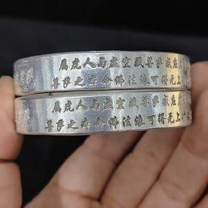 Puede incluir: Un brazalete de metal color plata que presenta dos bandas apiladas con un acabado liso y pulido. La superficie exterior de cada banda está grabada con intrincados caracteres chinos negros dispuestos en dos filas horizontales a lo largo del metal.