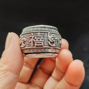 May include: A silver-toned metal ring featuring a wide, barrel-shaped band with an intricate mesh texture. The centre displays raised Chinese characters and decorative motifs, bordered by geometric patterns along the top and bottom edges.
