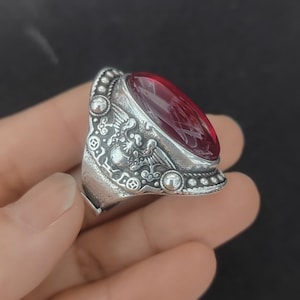 Puede incluir: Un anillo de tono plateado con una gran piedra cabujón de forma ovalada en color rojo intenso engastada en el centro. La banda de metal muestra intrincados detalles en relieve, incluyendo motivos alados y patrones circulares en los bordes.