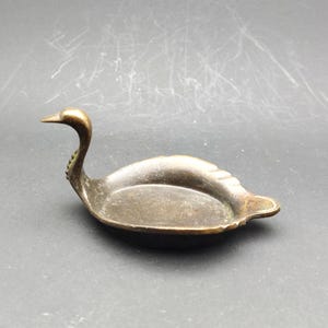 May include: A vintage brass swan-shaped dish. The swan has a long, curved neck and a small, detailed head. The dish is oval-shaped with a shallow bowl and a textured surface. The brass has a tarnished, antique finish.