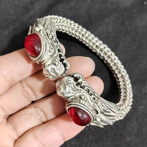 Puede incluir: Pulsera plateada con terminales de cabeza de dragón y ojos de piedras preciosas rojas. La pulsera tiene un diseño de cadena tejida. Las cabezas de dragón están intrincadamente detalladas y las piedras preciosas son de color rojo intenso.