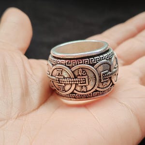 May include: A silver-toned metal ring featuring a repeating pattern of circular coin motifs with embossed Chinese characters and a decorative geometric border along the top and bottom edges. The band has a wide, cylindrical shape with a dark finish.