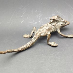 May include: A bronze-colored lizard figurine with detailed scales and a frill around its head. The reptile is posed in a walking position, with its legs extended. The sculpture is on a dark gray surface.