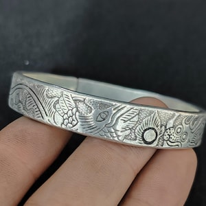 May include: A silver-toned metal cuff bracelet featuring an intricate embossed pattern of stylized fish, scales, and swirling wave-like motifs across the outer surface. The design has a matte, textured finish with darkened recessed areas.