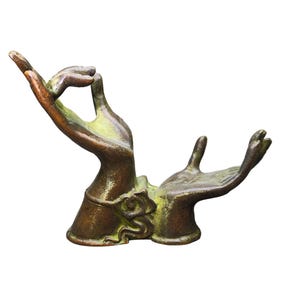 May include: A bronze-toned metal sculpture featuring two hands joined at the base. One hand is raised with fingers curled in a gesture, while the other hand is open and palm-up. The surface has a weathered, dark brown finish with green patina accents.