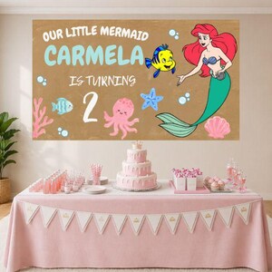 May include: A rectangular backdrop featuring a tan background with the text OUR LITTLE MERMAID CARMELA IS TURNING 2, illustrated with a red-haired mermaid, a yellow fish, a pink octopus, a blue starfish, and a pink seashell on a table.