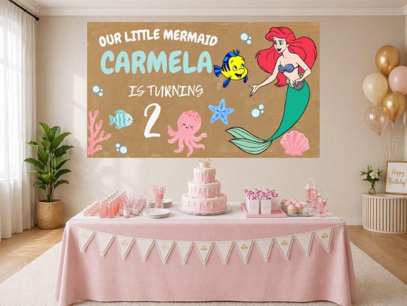 May include: A rectangular backdrop featuring a tan background with the text OUR LITTLE MERMAID CARMELA IS TURNING 2, illustrated with a red-haired mermaid, a yellow fish, a pink octopus, a blue starfish, and a pink seashell on a table.